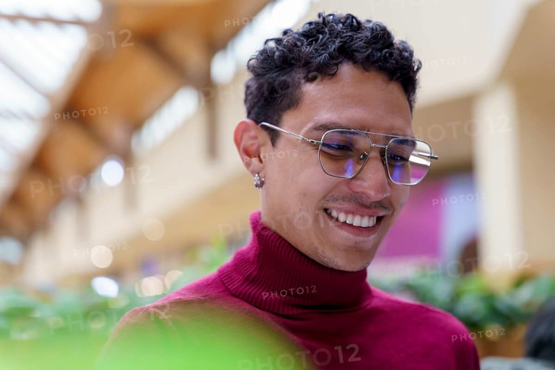 A happy young man from Gen Z smiles brightly in a public indoor space, showcasing a cheerful and modern lifestyle with a vibrant environment in the background
