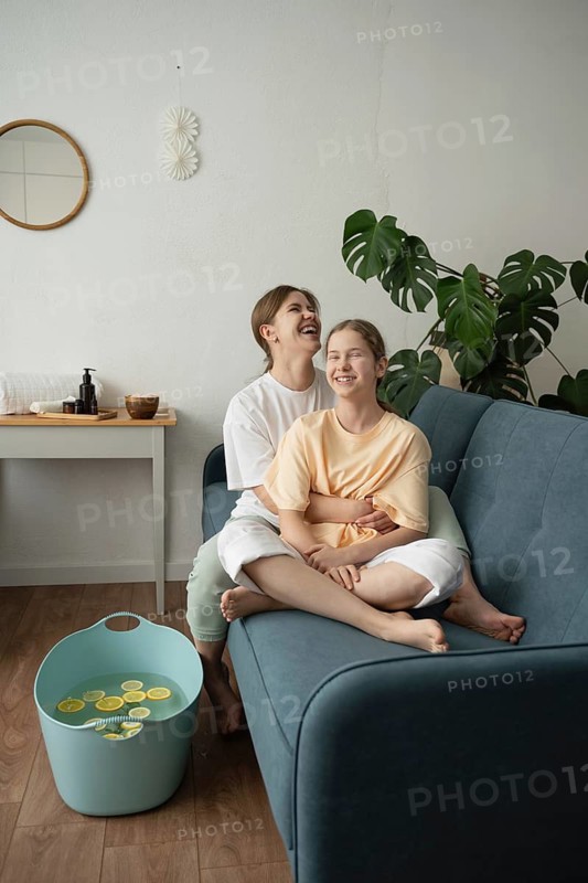 A mother and daughter share a joyful moment while engaging in a self-care routine at home. They are smiling and relaxed, sitting on a cozy sofa, surrounded by calming decor