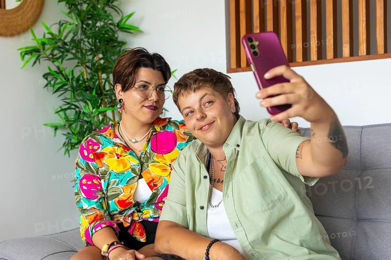 A cheerful lesbian couple sitting on a sofa, taking a selfie with a smartphone. The cozy living room setting features a potted plant and wooden decor, creating a relaxed atmosphere