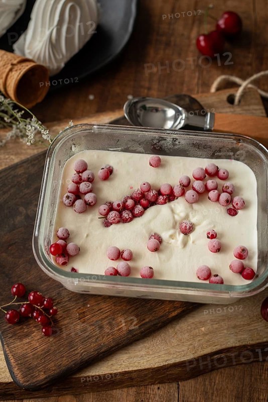 Top view of creamy vanilla ice cream sprinkled with frozen red currants in a rectangular glass dish, with fresh currants and a wooden background