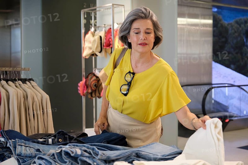 A mature woman in a yellow top explores clothing options at a fashion store. She is focused on selecting garments, showcasing a stylish and discerning shopping experience