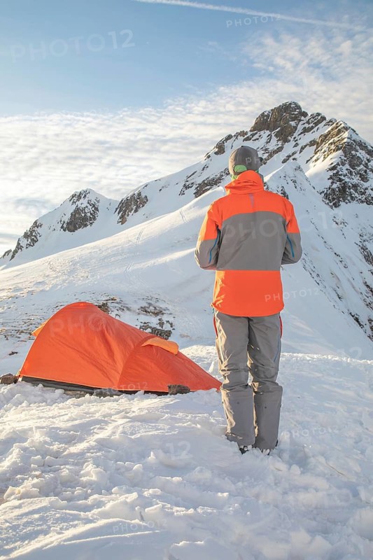 A person stands near a bright orange tent amidst snowy mountains under a clear sky. The scene captures the essence of solitude and adventure in a pristine winter landscape
