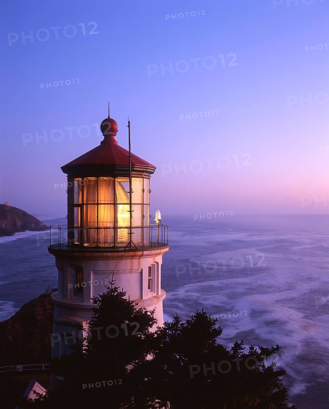 The Heceta Head Lighthouse glows warmly at dusk, standing against the vast Pacific Ocean near Florence, Oregon. The fading twilight casts a serene purple and pink hue over the coast