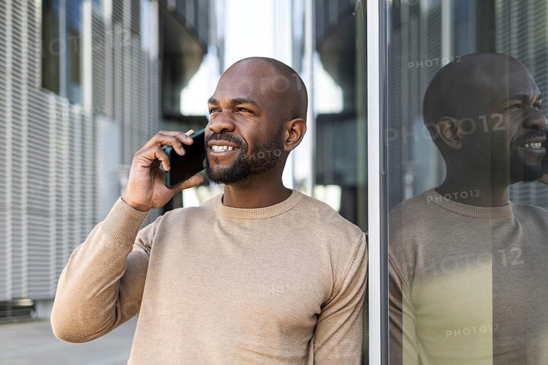 Smiling business cuban man in a beige sweater talks on a smartphone outside against a modern glass and steel backdrop, exemplifying urban communication and connectivity