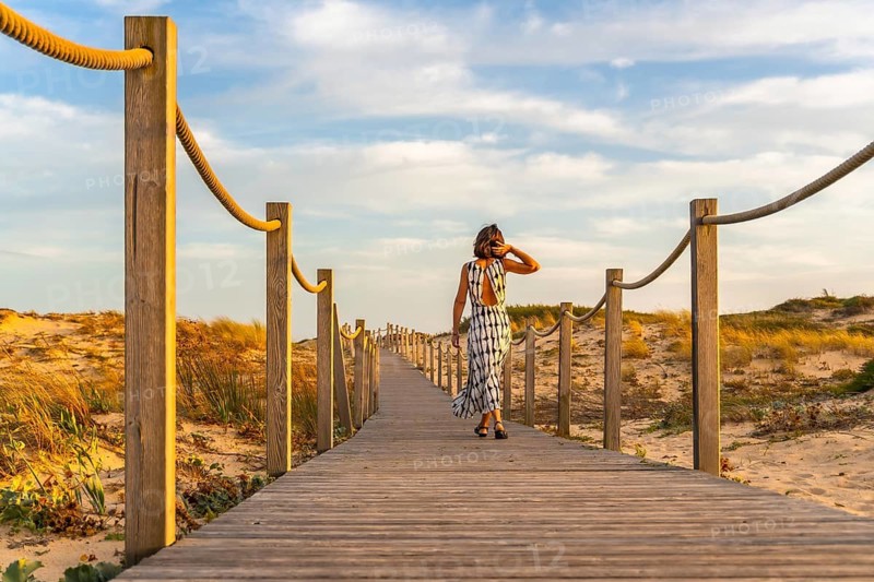 Woman in a dress enjoying a leisurely walk along a wooden boardwalk, leading through sand dunes to a beach under a warm, golden sunset sky, signifying summer holidays and peaceful moments