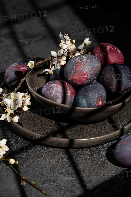Vibrant, naturally dyed Easter eggs rest in a dark ceramic bowl, adorned with delicate cherry blossoms, capturing the essence of spring