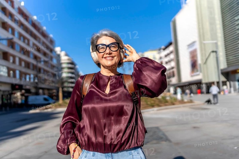 Senior woman strolling through a sunny city street, smiling and listening to music with headphones, backpack on, enjoying travel, independence and vibrant urban leisure