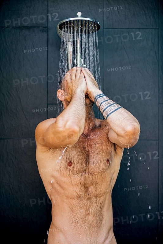 A man enjoys a refreshing shower under a modern showerhead, with water cascading over his body as he leans against a sleek, dark-tiled wall, conveying relaxation and hygiene