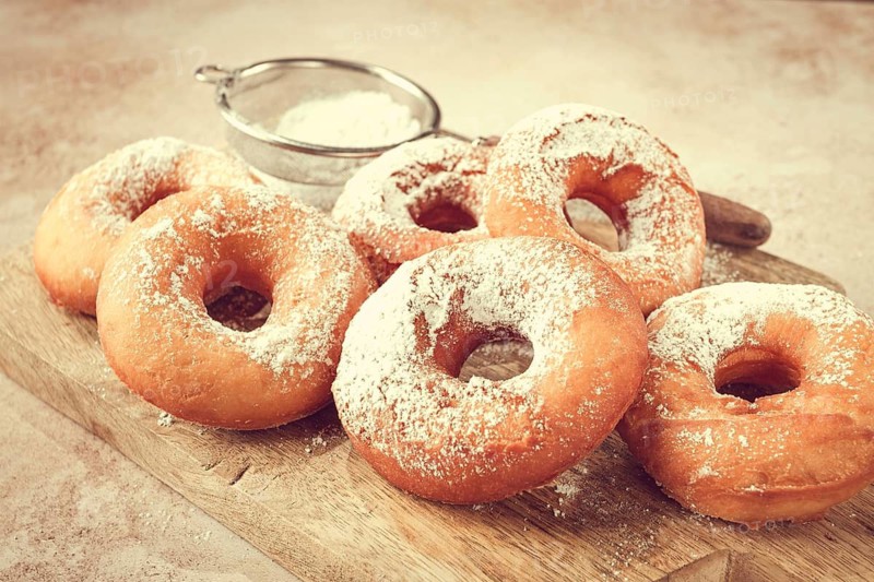 A wooden board holds several freshly made donuts. The donuts are round and have powdered sugar on top. A small sieve is nearby for additional sugar