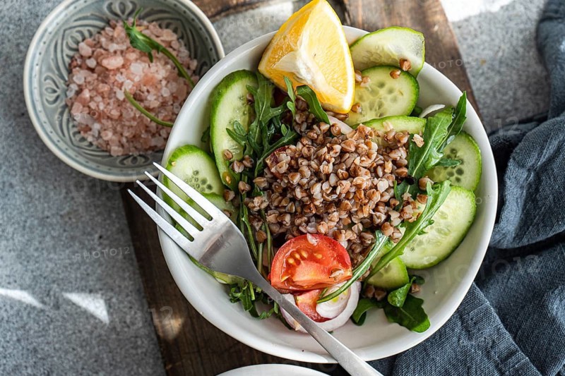 A nutritious lunch featuring fresh rocket, chopped oregano, cherry tomatoes, cucumber, Himalayan salt, red onion, steamed buckwheat, lemon, and organic olive oil in a bowl