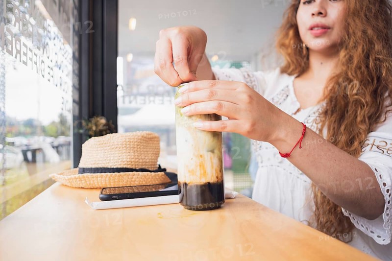 Young American Latin woman with long hair in a cozy cafe enjoying a drink. She wears a white top while sitting at a wooden table with sunlight streaming through the window