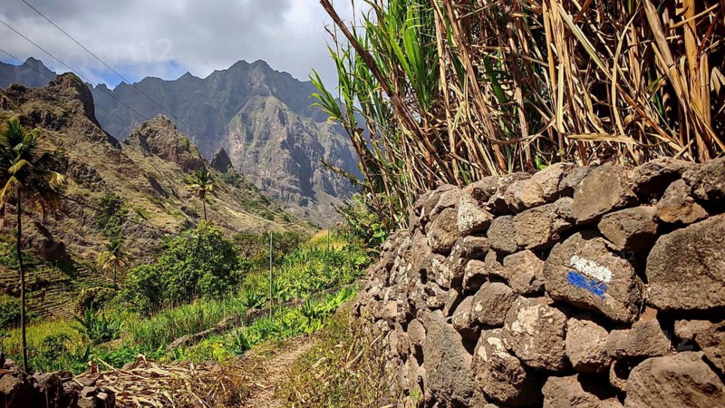 Stone wall with hiking trail, blue and white marker, behind it majestic mountains under cloudy sky, spectacular hiking, Santo Antao, Cabo Verde, Cape Verde, Cape Verde, Cape Verde