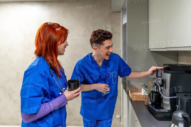 Two cheerful young medical professionals in blue scrubs enjoying a coffee break, conversing and operating the coffee machine while sharing a moment of relaxed camaraderie in a hospital setting