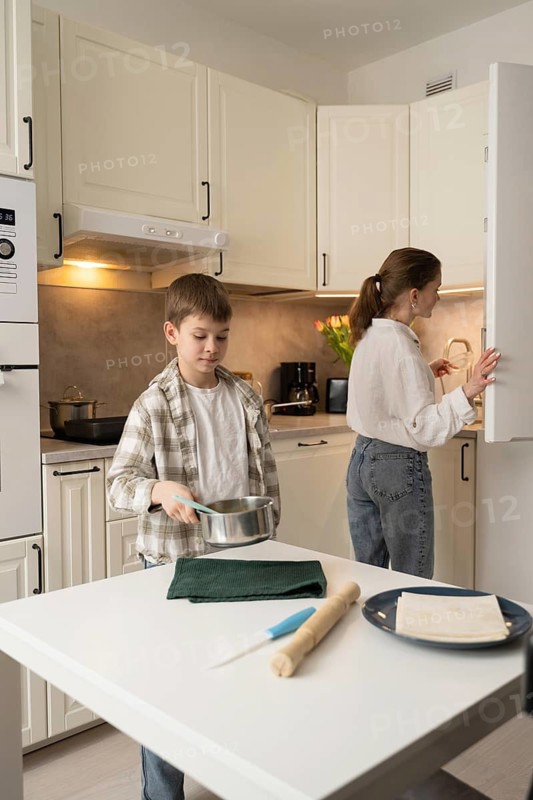 A mother and child engage in cooking activities in a cozy kitchen. The boy holds a pot while the mother checks the fridge. The scene reflects teamwork and family bonding