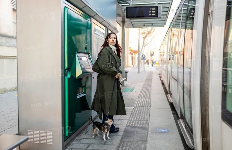 A woman with a small chihuahua waits near a tram station, standing next to a ticket machine. The urban setting captures modern city life and public transport ambiance