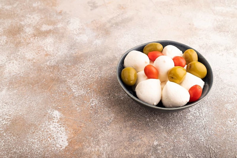 White Mozzarella cheese, with tomatoes and olives in blue bowl on brown concrete background, side view, copy space