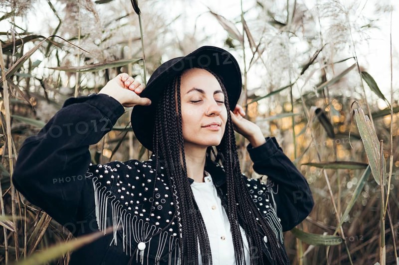 A woman with braids stands serenely in a field at sunset, wearing a stylish hat and closing her eyes to embrace the tranquil atmosphere around her