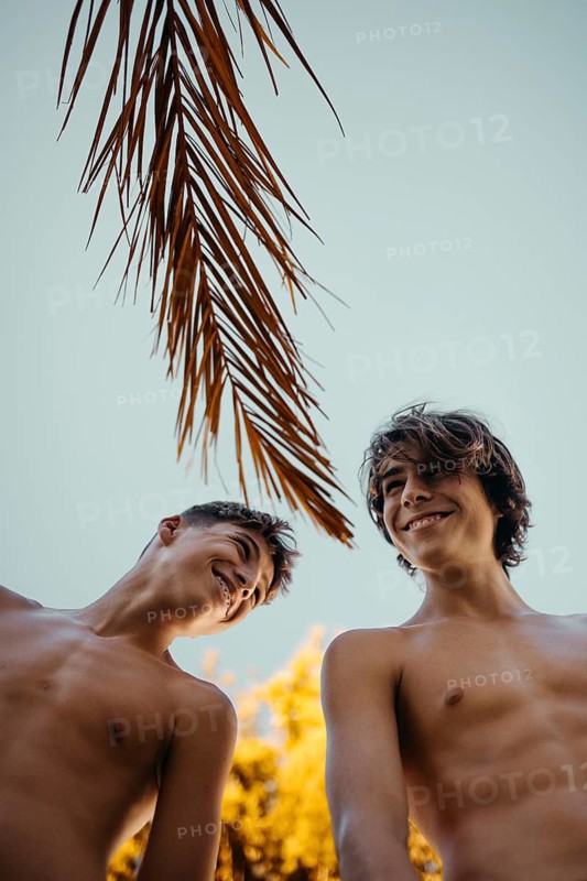 Two teen boys enjoy a sunny day outdoors, laughing under palm tree leaves against a clear sky. Their carefree expressions capture the joy of friendship and summer fun