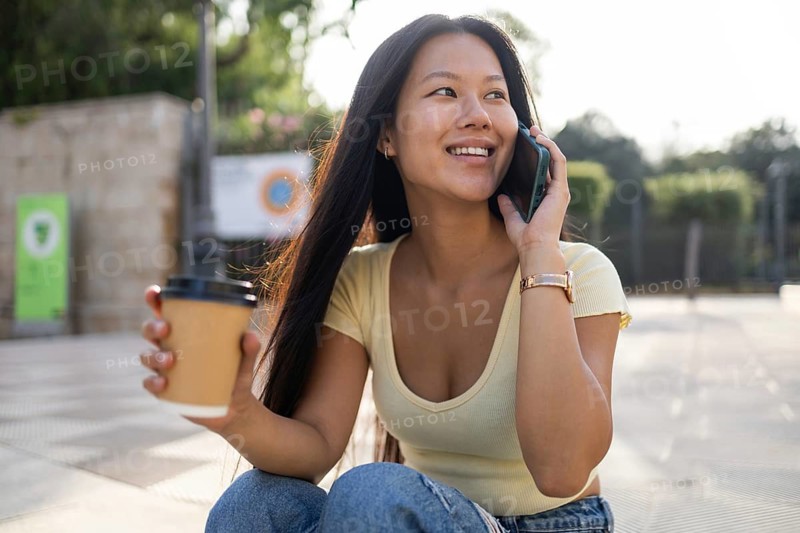 A young Chinese woman enjoys a pleasant summer day in the city, engaging in a cheerful conversation on her phone while holding a cup of coffee, surrounded by urban greenery