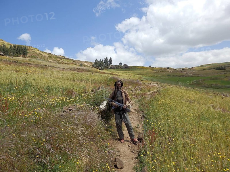 Man standing smiling with a rifle, Kalashnikov in a flower-rich field surrounded by rolling hills, Simien National Park, Ethiopia