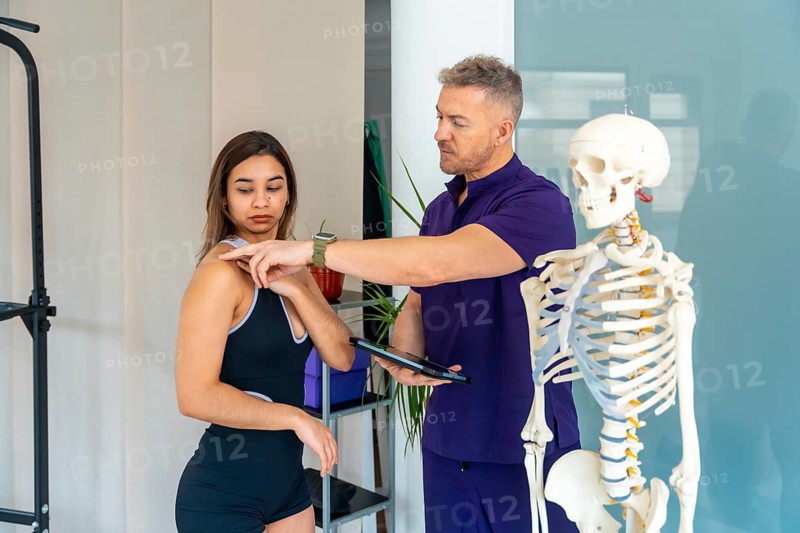 Physiotherapist showing a female patient shoulder points on her body while discussing a treatment plan in a clinic, with a skeleton model next to them