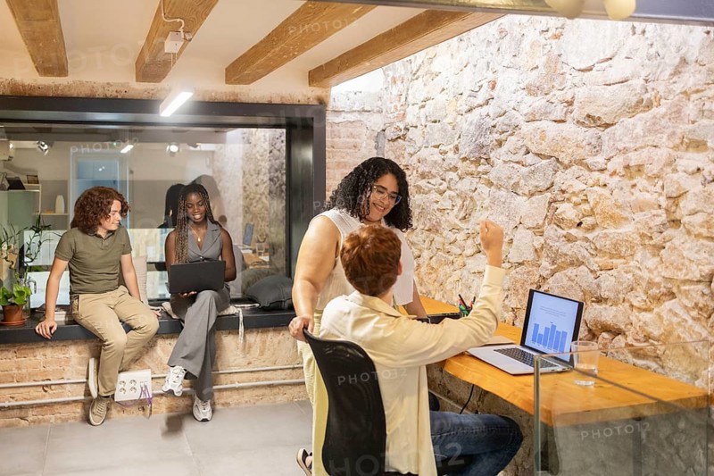 A diverse group of coworkers in a stylish office, discussing ideas. Two are using laptops, engaging in teamwork. The environment is inclusive and tech-friendly