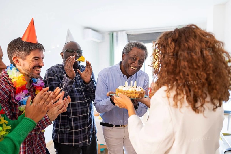 A group of people celebrates a birthday with smiles and clapping. A person presents a festive cake with lit candles, creating a joyful and heartwarming atmosphere