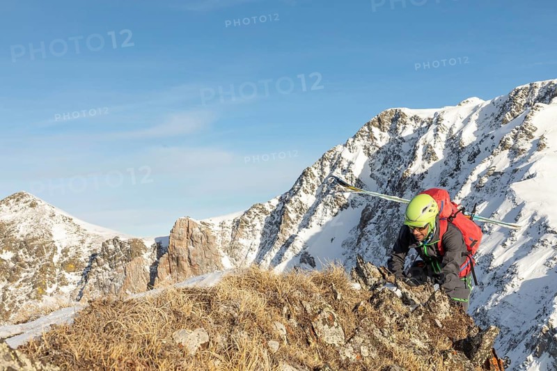 A ski tourer in colorful gear climbs a rugged, snowy mountain ridge under a bright blue sky. The scene captures the thrill and challenge of alpine adventure