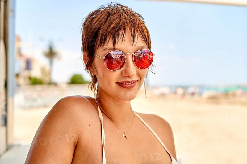 A woman with short hair relaxes on a sunny beach wearing round sunglasses and a bikini. The vibrant atmosphere captures the essence of summer fun by the sea