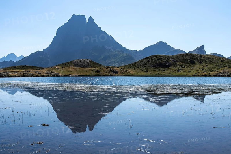 A breathtaking view of Midi d'Ossau in the French Pyrenees, reflected in a serene mountain lake The clear blue sky and lush green hills enhance the scenic beauty