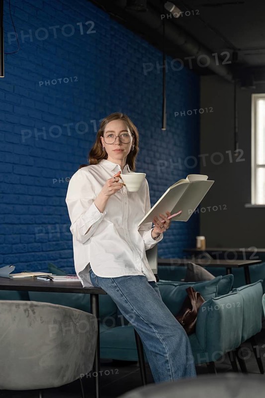 A woman in a white shirt and jeans enjoys a cup of tea while reading a book in a modern cafe with blue brick walls and chic furniture. The setting is cozy and tranquil