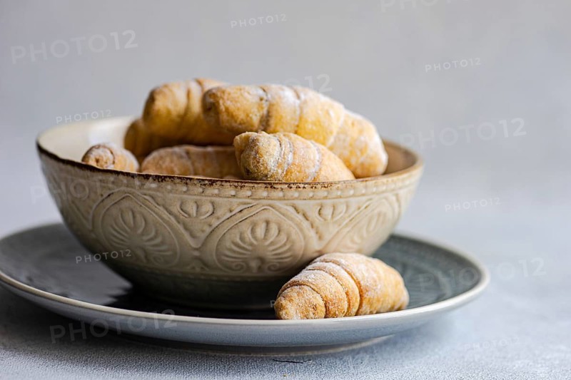From above, a rustic ceramic bowl filled with freshly baked homemade croissants is placed on a matching plate, showcasing the delicious pastries dusted with powdered sugar