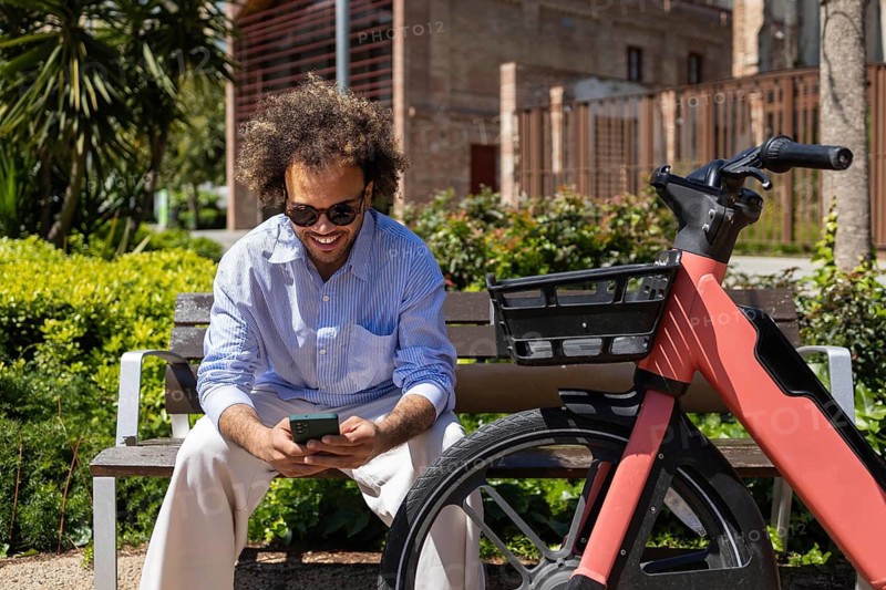 Afro man sitting on a park bench, smiling while looking at his smartphone. A bicycle is parked beside him. Capturing a moment of leisure and connectivity in an urban setting