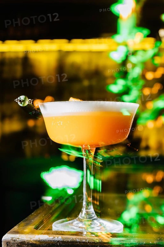 A vibrant peach cocktail with a frothy top, elegantly served in a coupe glass. The cocktail sits on a reflective glass bar, illuminated by warm golden lighting and a green ambiance