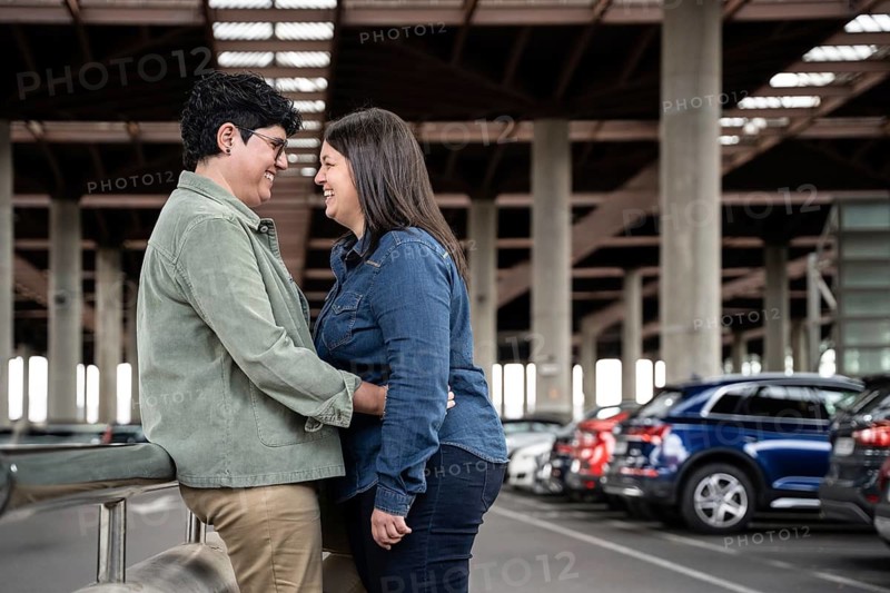 A joyful curvy lesbian couple embraces in an urban parking area. The moment captures a sense of connection and happiness amidst the city backdrop of cars and structures