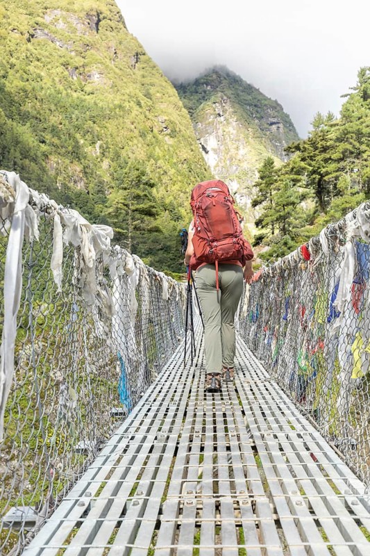 A woman with a red backpack treks across a suspension bridge surrounded by stunning mountain scenery on an Everest trail, showcasing adventure and outdoor exploration