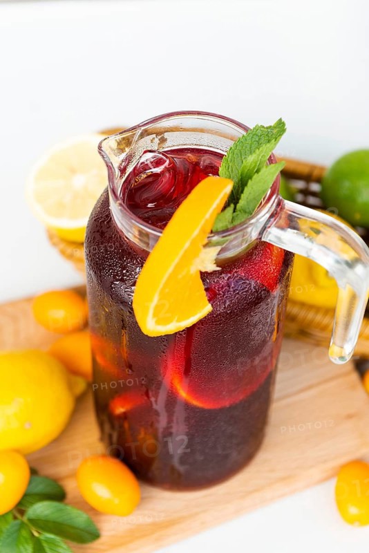 A glass pitcher of sangria filled with citrus slices, berries, and ice, garnished with mint leaves, sitting on a wooden board surrounded by fresh lemons and kumquats
