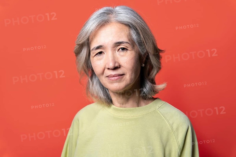 Portrait of an Asian senior woman with gray hair, wearing a light green sweater. She stands confidently against a vibrant red background, exuding wisdom and grace