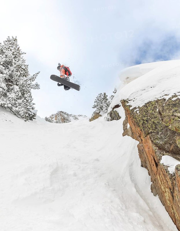 A snowboarder performs an airborne trick over a rocky cliff, surrounded by snow-covered trees and landscape, showcasing dynamic motion and winter sports athleticism