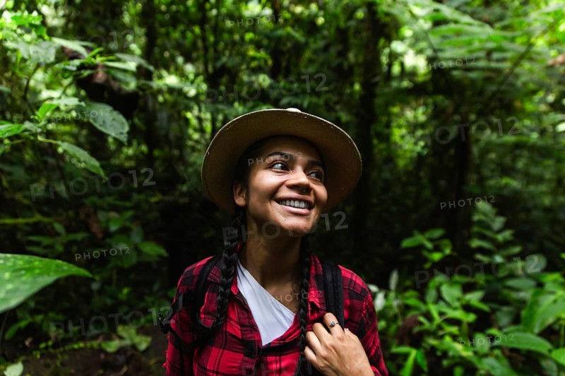 An Ecuadorian woman, dressed in a red plaid shirt and sporting a hiking backpack, smiles joyfully as she explores the dense, green Choco Andino rainforest in Ecuador. This image captures her engaging with the verdant environment