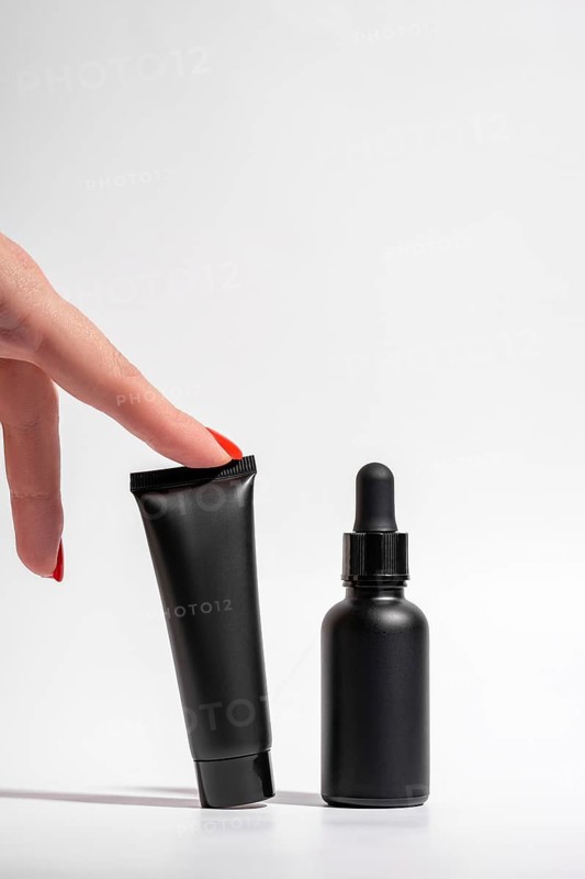 Cropped unrecognizable female hand with red manicure points at black cosmetic packaging against a clean white backdrop