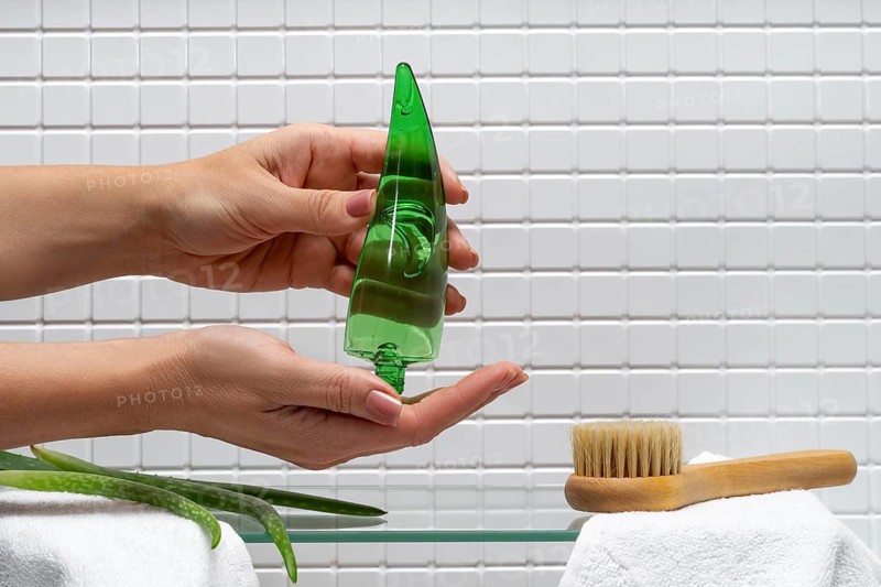 Cropped unrecognizable woman dispenses aloe vera gel onto her hand against a tiled bathroom background with body care essentials visible