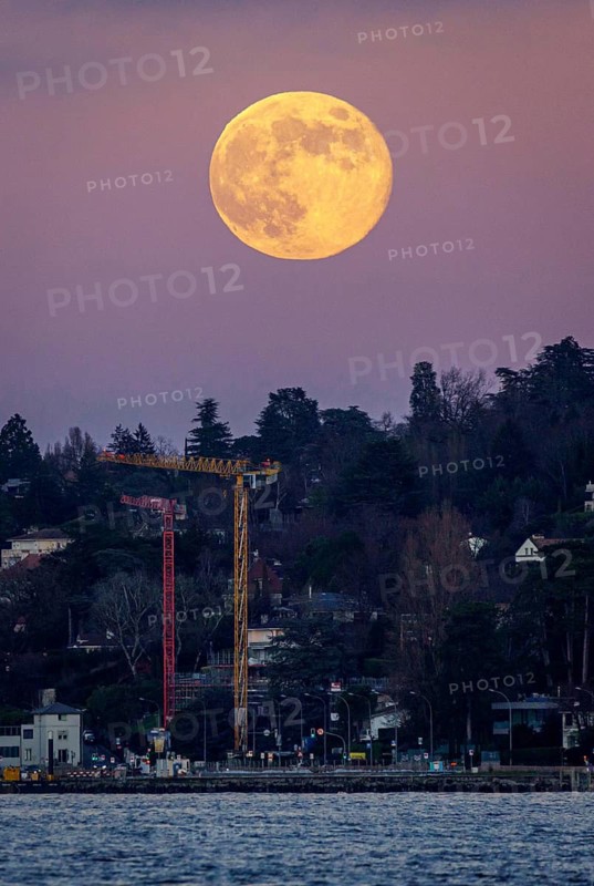 A vibrant full moon rises against a serene evening sky in Geneva, casting a warm glow over an urban landscape with a construction crane and lush trees by the waterfront