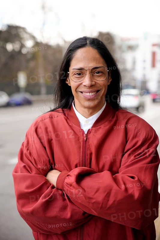 A man in a red jacket stands confidently on a city street, smiling warmly with arms crossed. The background features a soft focus urban landscape and parked cars
