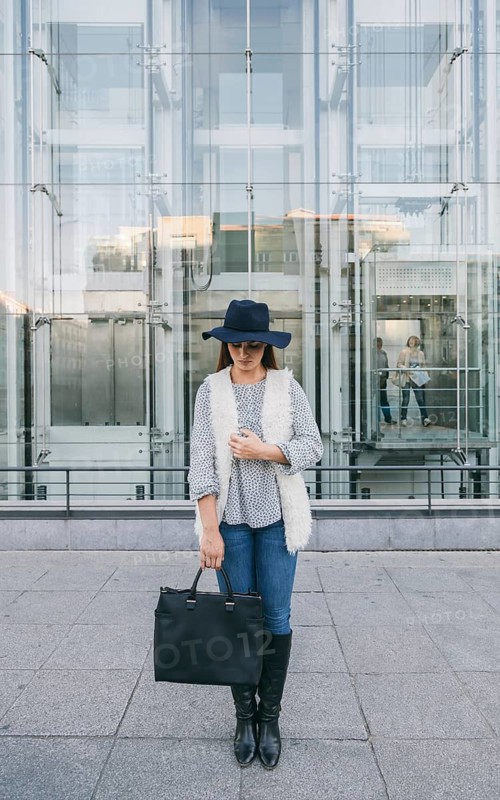A fashionable woman stands in front of a modern glass building wearing a wide-brimmed hat, blue jeans, and a white vest. She holds a large black handbag and boots complete her chic look