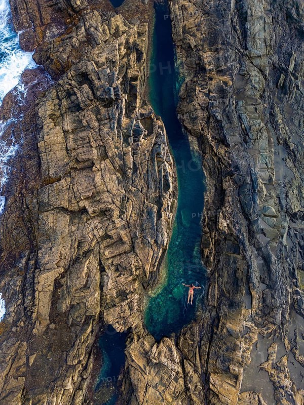 Top view of an unrecognizable woman floating peacefully in a natural pool amid rugged cliffs at Tindaya Beach, Fuerteventura. This aerial drone image captures the tranquility and untouched nature of the location