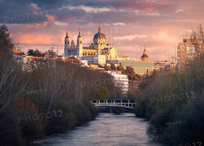 Captivating sunset view of the majestic Almudena Cathedral of Madrid bathed in warm hues, set against a dramatic sky. The Manzanares river and surrounding architecture add to the serene atmosphere