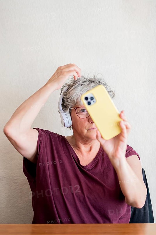 A senior woman with headphones uses a yellow smartphone for a selfie. She wears glasses and a burgundy shirt, capturing a candid moment in a relaxed setting