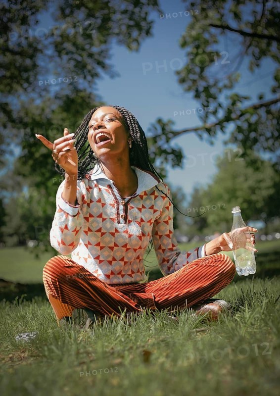 A cheerful woman sits on the grass, embracing the beauty of nature with a refreshing bottle of water. The vibrant outdoor setting enhances her joyful expression