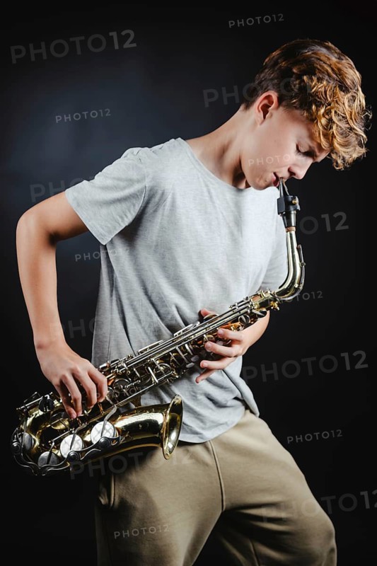 A teenage boy passionately playing a saxophone in a dimly lit studio, highlighted against a black background, showcasing concentration and musical expertise